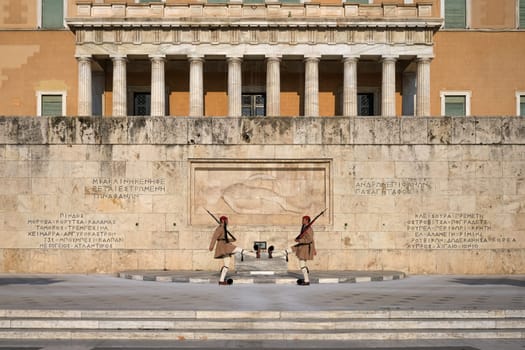 Ceremony of Presidential guard Evzones by the Greek tomb of Unknown Soldier. Evzones in summer uniform face each other, legs up. Parliament building. Wide shot.