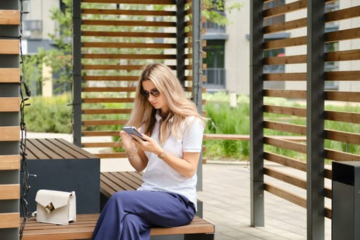 Woman in polo shirt and trousers sitting on bench with smartphone