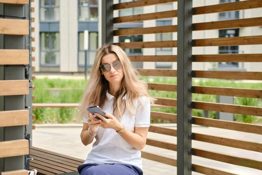 Woman in white shirt and sunglasses on bench with smartphone