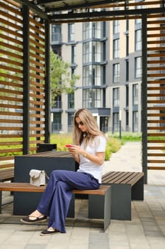 Woman in polo shirt and trousers sitting in gazebo on street with smartphone
