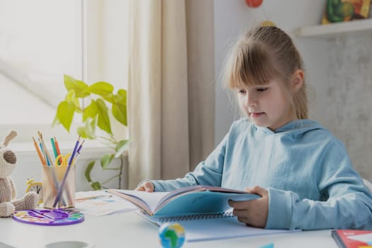 Schooler cute little girl sitting at desk in bedroom, holding a book and reading literature, doing homework, kid studying at home, getting ready before exams. Home education, homeschooling