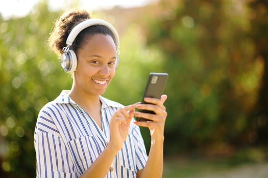 Happy black woman wearing headphone and holding smart phone posing looking at camera listening to music in a park