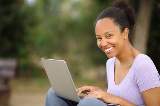 Happy black woman using laptop looking at you sitting in a park