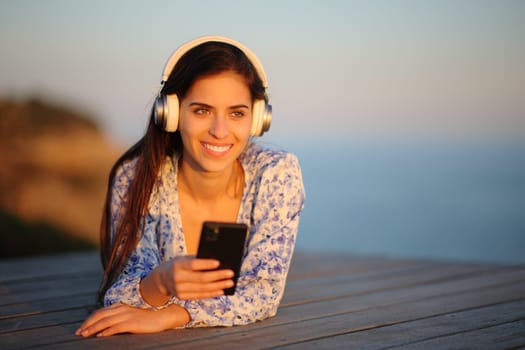 Happy woman wearing headphone lying at sunset listening to music on phone on the beach and contemplating