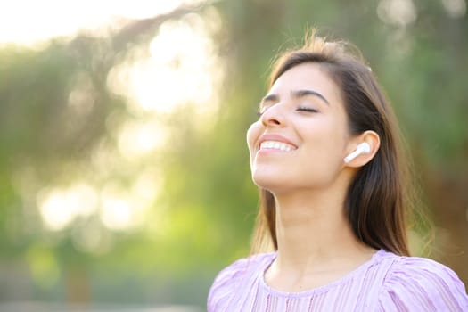 Happy woman breathing fresh air wearing earbud in a green park