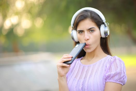 Suspicious woman looking at you listening digital audio with headphone and smartphone in a park