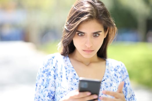 Front view portrait of a perplexed woman checking phone surprising news in the street