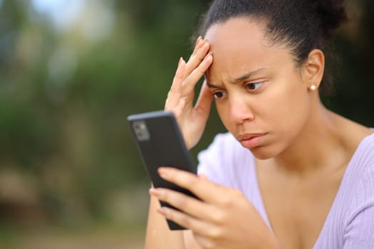 Worried black woman checking bad news on cell phone in a park