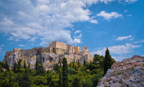 View of Acropolis hill from Areopagus hill in summer daylight with great clouds in blue sky, Athens, Greece. UNESCO world heritage. Propylaea entrance, Agrippa's monument, Parthenon. Fresh green foliage and stones and rocks of hilltop