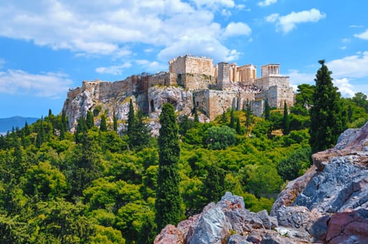 View of Acropolis hill from Areopagus hill in summer daylight with great clouds in blue sky, Athens, Greece. UNESCO world heritage. Propylaea entrance, Agrippa's monument, Parthenon. Fresh green foliage and stones and rocks of hilltop