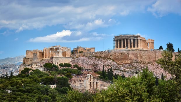 View of Acropolis hill with Propylaea and Parthenon and theater of Odeon below it in Athens, Greece from the hill of Philoppapos or Muses in summer daylight with great clouds in blue sky. Great fresh foliage of olives and cedar trees. Close view