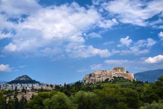 View of Acropolis hill with Propylaea and Parthenon and hill of Lycabettus in background in Athens, Greece from Pnyx hill in summer daylight with great clouds in blue sky. Great fresh foliage of olives and cedar trees in foreground