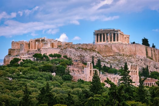 View of Acropolis hill with Propylaea and Parthenon and theater of Odeon below it in Athens, Greece from the hill of Philoppapos or Muses in summer daylight with great clouds in blue sky. Great fresh foliage of olives and cedar trees. Close view