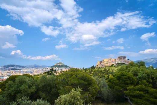 View of Acropolis hill with Propylaea and Parthenon and hill of Lycabettus in background in Athens, Greece from Pnyx hill in summer daylight with great clouds in blue sky. Great fresh foliage of olives and cedar trees in foreground