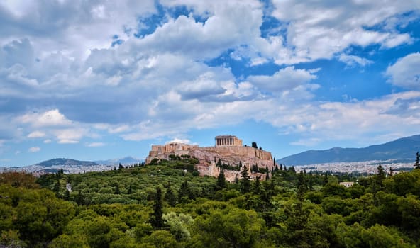 View of Acropolis hill with Propylaea and Parthenon and theater of Odeon below it in Athens, Greece from the hill of Philoppapos or Muses in summer daylight with great clouds in blue sky. Great fresh foliage of olives and cedar trees. Wide shot