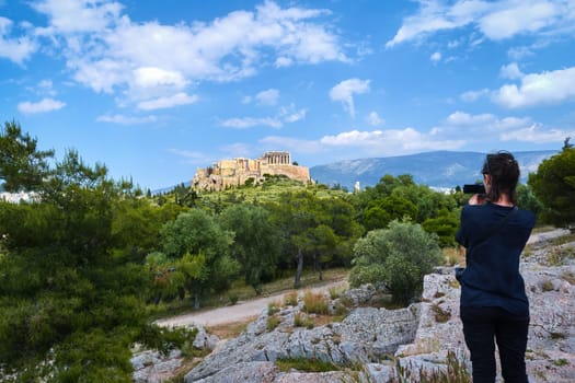 Unrecognizable female tourist dressed in black shoots a view of Acropolis hill with Parthenon and Lycabettus hill in background, Athens, Greece from Pnyx hill on summer day with great clouds in blue sky. Great fresh foliage of olives and cedar