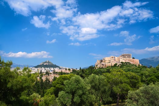 View of Acropolis hill with Propylaea and Parthenon and hill of Lycabettus in background in Athens, Greece from Pnyx hill in summer daylight with great clouds in blue sky. Great fresh foliage of olives and cedar trees in foreground