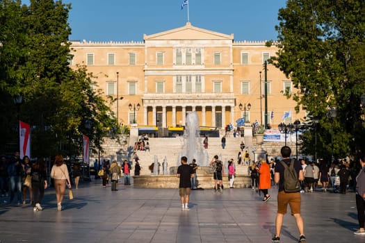 Citizens and tourists roaming in Syntagma square in afternoon. View of Hellenic Parliament and Syntagma lit by bright afternoon sun. Center of Athens, Greece, iconic square, tourist attraction, patriotic symbol for Greek nation.