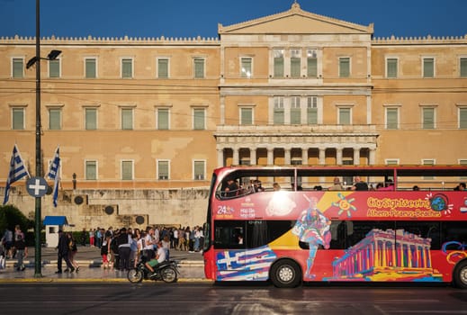 Tourists, city traffic, Hop-on-Hop-off tourist bus in Syntagma square and Hellenic Parliament building lit by afternoon sun. Tourist and patriotic center of Athens, Greece, daily ceremony of changing guards i.e. evzones in soft summer sunlight.