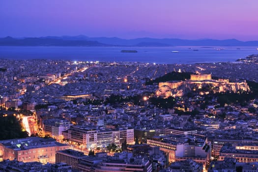 Panoramic wide night view of Athens and Acropolis shot from Lycabettus hill at dusk. Parthenon and Erechtheion lit up by superb night lights. Saronic Gulf and Hellenic Parliament. Famous iconic view of UNESCO world heritage site. After sunset