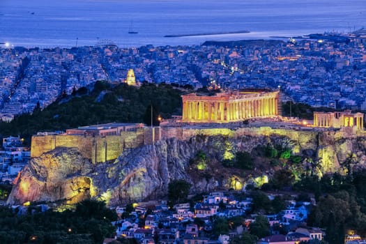 Close view of Acropolis hill with Parthenon and Erechtheion lit up by superb night lights and Philoppapos monument in distance. Night lights of Athens and its harbour. Famous iconic view of UNESCO world heritage site.