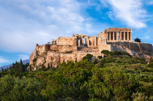 View of Acropolis hill with Propylaea and Parthenon in Athens, Greece from Pnyx hill in summer afternoon with great clouds in blue sky. Great fresh foliage of olives and cedar trees in foreground. Close shot. Awesome composition