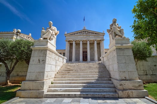 Marble statues of Plato and Socrates, ancient Greek philosophers, seated in chairs, main entrance to Academy of Athens, national highest research center of Greece. Neoclassical architecture. Bright summer day, majestic blue sky. Greek national flag