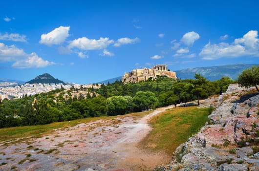 View of Acropolis hill with Propylaea and Parthenon and hill of Lycabettus in background in Athens, Greece from Pnyx hill in summer daylight with great clouds in blue sky. Great fresh foliage of olives and cedar trees in foreground