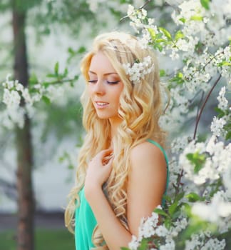 Beautiful young woman with blonde long hair in spring blooming garden on a sunny day against flowers trees background