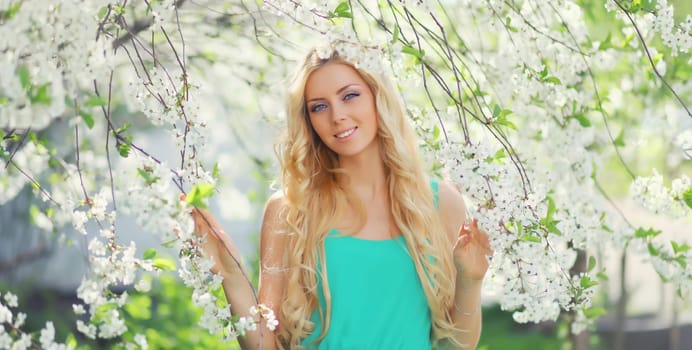 Beautiful young woman with blonde long hair in spring blooming garden on a sunny day against flowers trees background