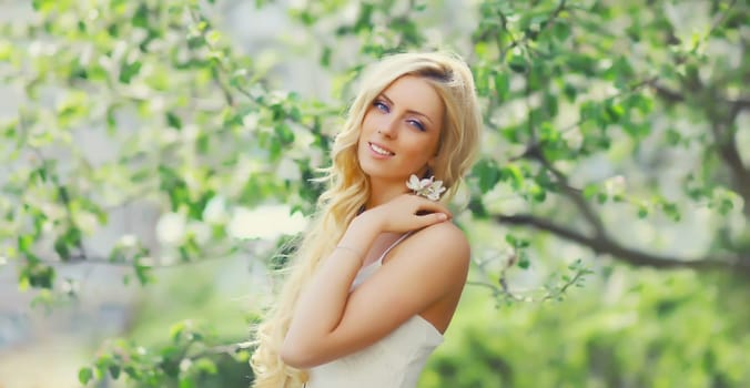 Beautiful young woman with blonde long hair in spring blooming garden on a sunny day against flowers trees background