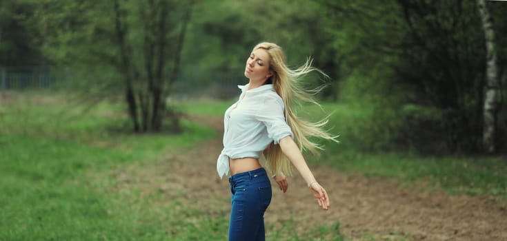 Beautiful young woman with windblown blonde long hair enjoying fresh air, wind on a summer day in forest against green trees background