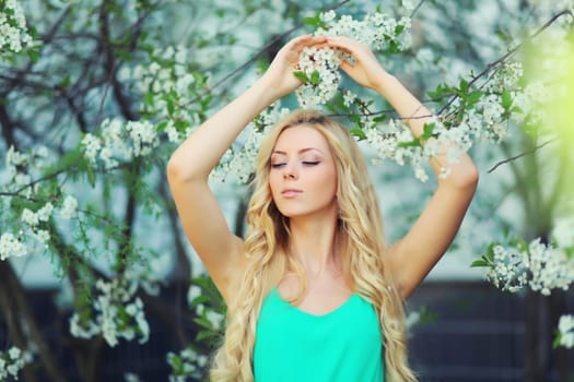 Beautiful young woman with blonde long hair in spring blooming garden on a sunny day against flowers trees background
