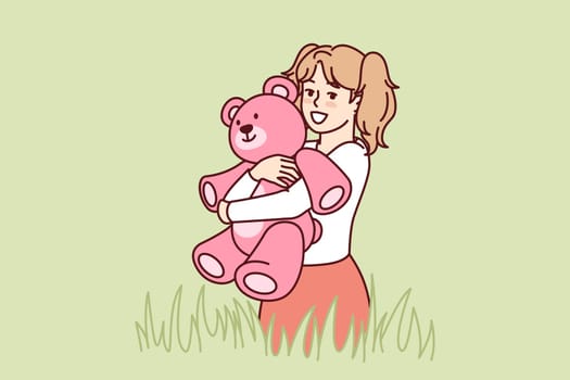 Happy little girl with teddy bear in hands stands in meadow among tall grass and looks at screen. Child with smile hugs beloved toy bear, presented for birthday, during summer walk