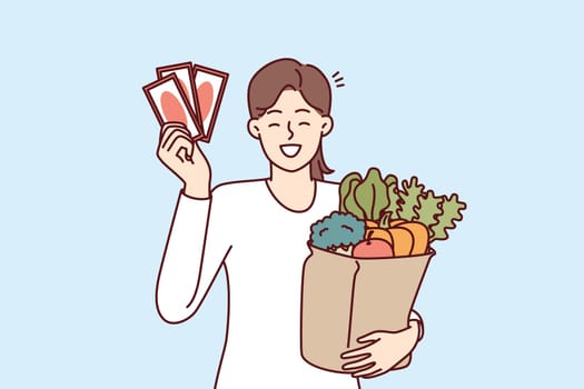 Woman shopper holding gift vouchers from supermarket and paper bag filled with fresh vegetables. Happy girl showing off discount coupons or vouchers for shopping at grocery store at bargain prices
