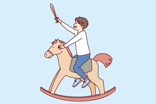 Cheerful boy sits astride toy horse and holds knight sword in hand dreaming about saving princess. Positive boy on steed-rocker enjoys vintage toys that give opportunity to dream up