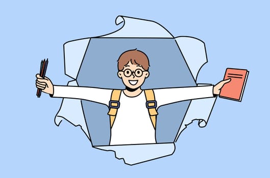 Little schoolboy with books and pencils rejoices at beginning of school year, looking out of hole in paper. Happy boy first grade student with backpack smiling for back to school concept