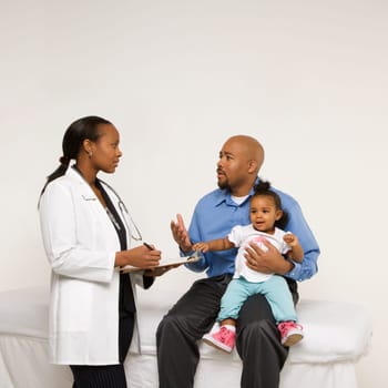 African-American father holding baby girl talking to female pediatrician.
