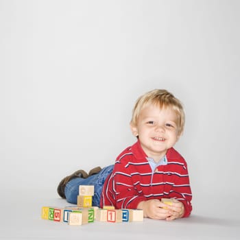 Studio portrait of Caucasian boy lying on stomach propped up on elbows playing with toy blocks.