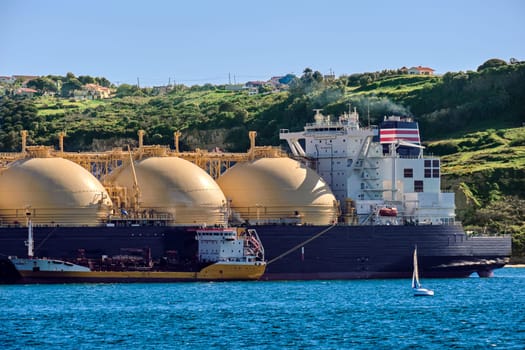 LNG or liquified natural gas tanker moored by industrial pier and green hill on sunny day. Alternative gas supply, commercial freight, energy crisis, regasification, diversification, fuel and energy generation.