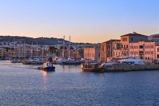 Fishing boat leaving Old Venetian harbour of Chania, Crete, Greece at sunrise. Sailing boats, yachts, tourist cruises anchored by piers. Grand Arsenal and Old Venetian shipyards known as Arsenali Veneziani or Neoria lit by first sunrays.