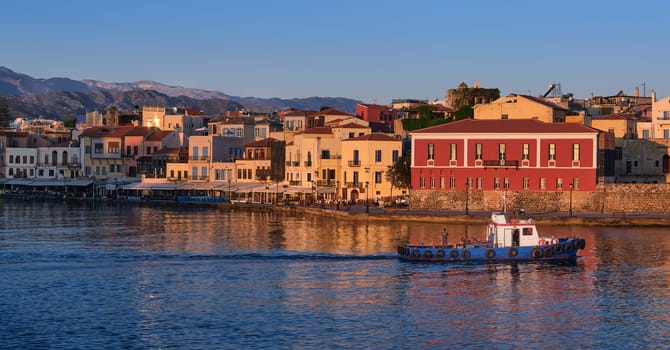Fishing boat passing by Old Venetian harbour promenade and Maritime museum of Crete in Chania, Crete, Greece, lit by first sunrays. Sunrise view of town and Cretan hills and mountains behind it.