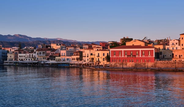 Sunrise view of Old Venetian harbour of Chania, Crete, Greece, shops, hotels, cafes and restaurants on its promenade. Maritime museum of Crete lit by first sunrays. Cretan hills and mountains in background.