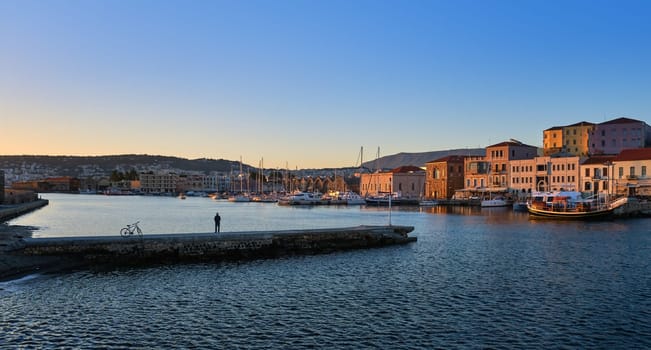 Sunrise in Old Venetian harbour of Chania, Crete, Greece. Grand Arsenal and Old Venetian shipyards or Arsenali Veneziani or Neoria. Sailing boats, yachts anchored by piers. Unrecognizable male and its bicycle standing on pier and enjoying the view