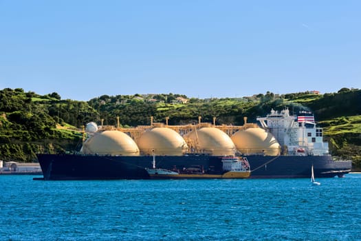 LNG or liquified natural gas tanker moored by industrial pier and green hill on sunny day. Alternative gas supply, commercial freight, energy crisis, regasification, diversification, fuel and energy generation.