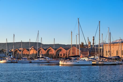 Sunrise over Old Venetian port and harbour of Chania, Crete, Greece. Tourist sailing boats, yachts anchored by pier and Old Venetian shipyards or Arsenali Veneziani or Neoria. Bell tower and minaret of Agios Nikolaos church, distant Cretan mountains