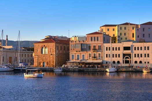 Fishing boat passing by Grand Arsenal and cafes and hotels of Old Venetian harbour quay of Chania, Crete, Greece, lit by first sunrays. Sunrise view of promenade and sailing boats anchored by piers.