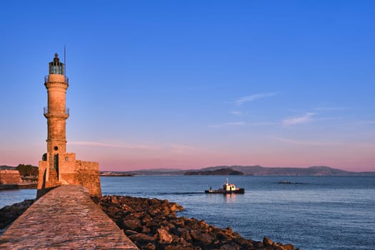 Fishing boat passing by Egyptian Lighthouse lit by first sunrays and leaving Old Venetian harbour of Chania, Crete, Greece at sunrise. Soft early morning colors of sky and Cretan hills and mountains in distance.