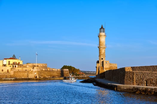 Fishing boat passing by famous Lighthouse and walls of old port, Firka fortress of Old Venetian harbour of Chania, Crete, Greece in early morning. Golden hour shot. Clear blue sky and sea.