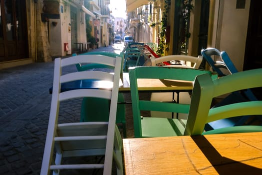 Early morning in empty narrow streets of old mediterranean town. Bright sunlight. Colorful chairs and tables in cafes and restaurants not ready. Selective focus of street furniture in foreground. Chania, Crete, Greece